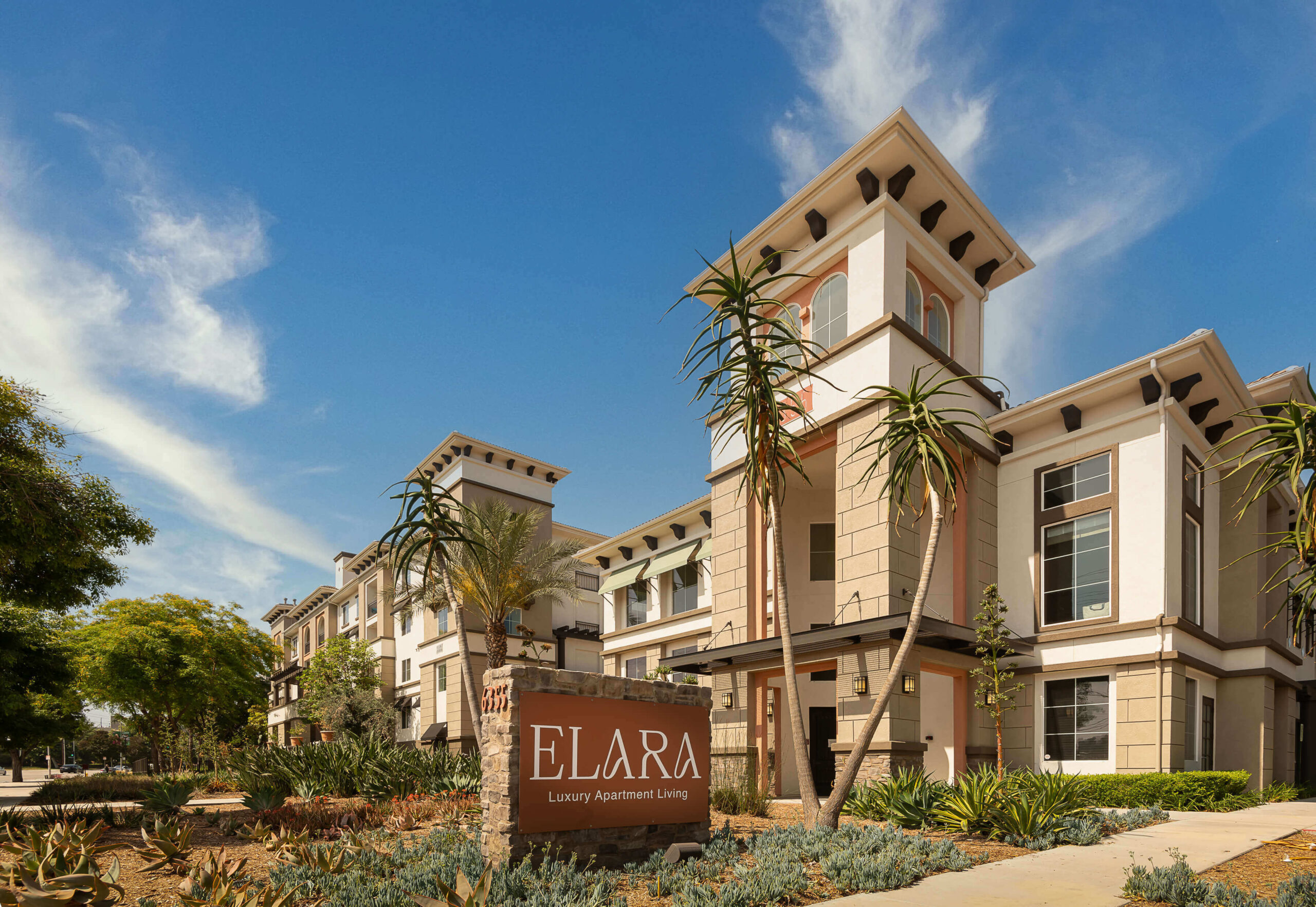 last photo homepage Woodland Hills, CA, Pet-Friendly Apartments - Elara - Leasing Office Building with Property Signage, Manicured Shrubs, Trees, and Blue Skies.